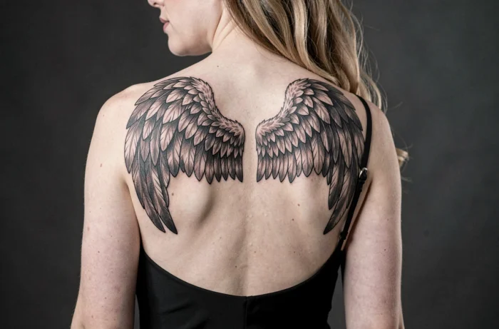 Tatouage ailes d'ange en micro-réalisme noir et gris sur le haut du dos d'une femme portant une robe noire dos nu