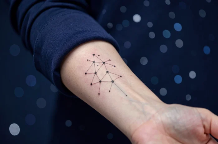 Tatouage constellation personnelle en points et lignes fines sur l'avant-bras d'une femme portant un haut bleu marine